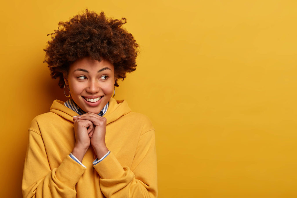 Portrait d'une femme à l'air agréable avec des cheveux bouclés, regardant avec joie de côté, étant dans un esprit élevé, habillée d'un sweat-shirt, pose contre un mur jaune. concept d'expressions et d'émotions du visage humain.